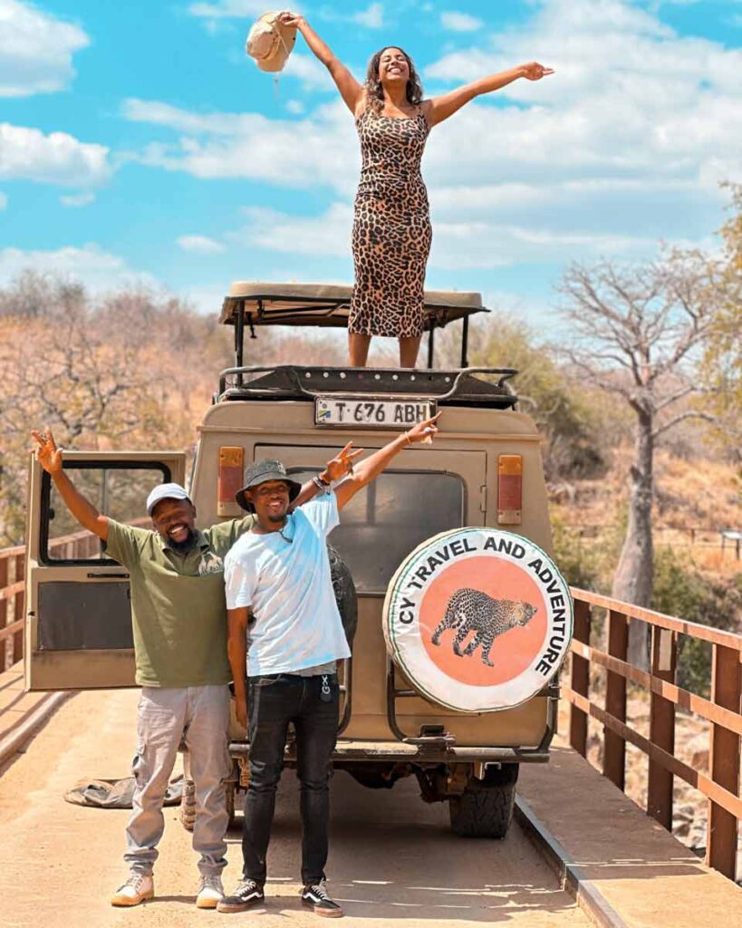 Two men posing in front of a safari jeep. A woman stands up on the roof ot the jeep smiling, with her hands in the air.