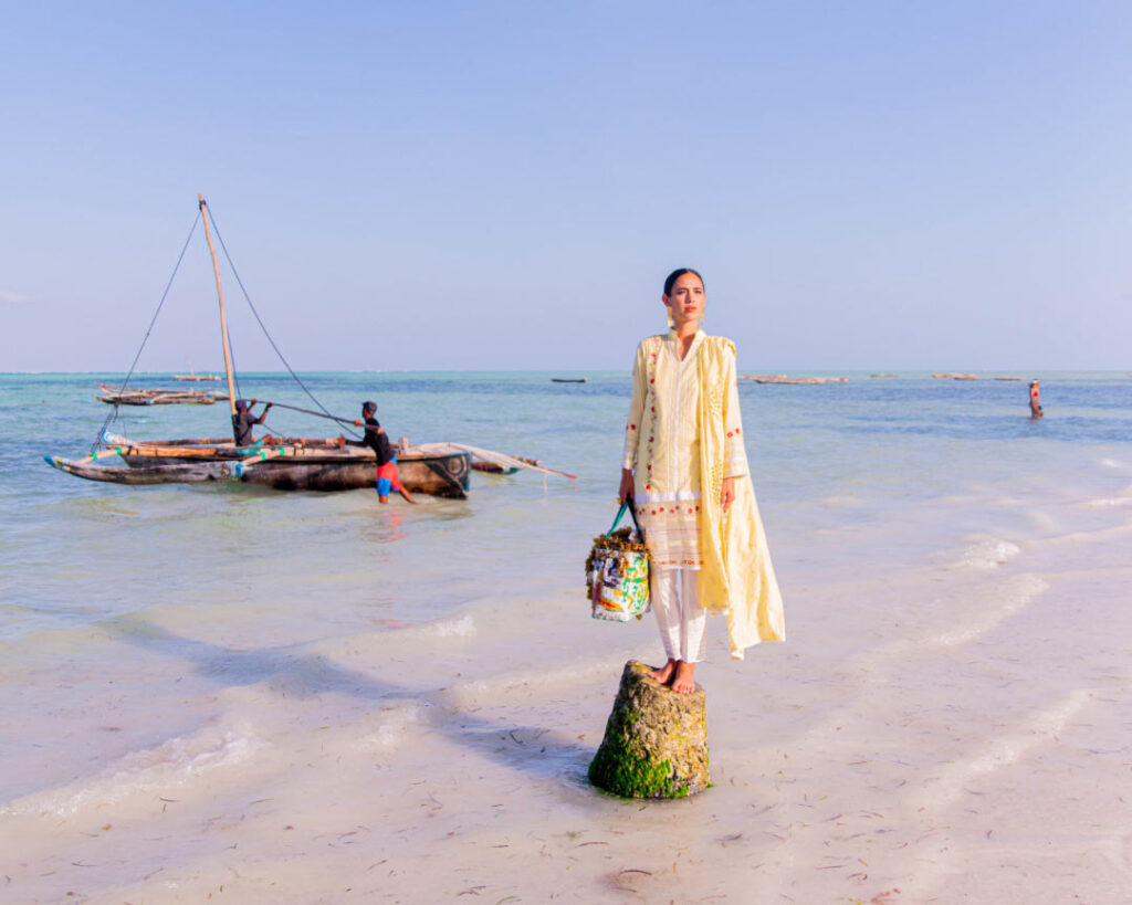A white woman standing on a rock, wearing a long ethnic yellow dress with red dots. She wears red lipstick. Behind her, there is the ocean. Two men are pushing a traditional Zanzibari fishing boat towards deeper water.
